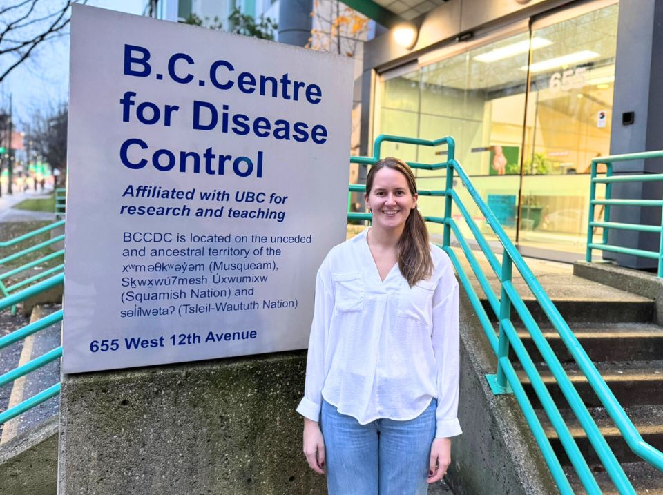 A woman in a white long-sleeved shirt stands smiling in front of a sign that says B.C. Centre for Disease Control