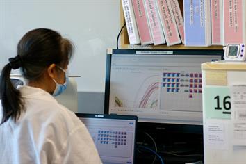 A lab technician in a white gown looks at a screen displaying wastewater surveillance data