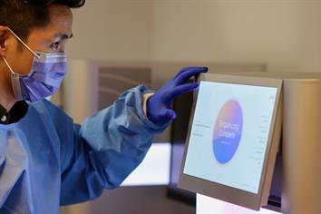 A person in a lab coat and a surgical mask looks at a screen that reads "Sequencing Complete"