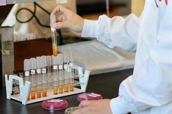 A lab technician in a white gown holds a sealed test tube with a brown and yellow wastewater sample inside of it